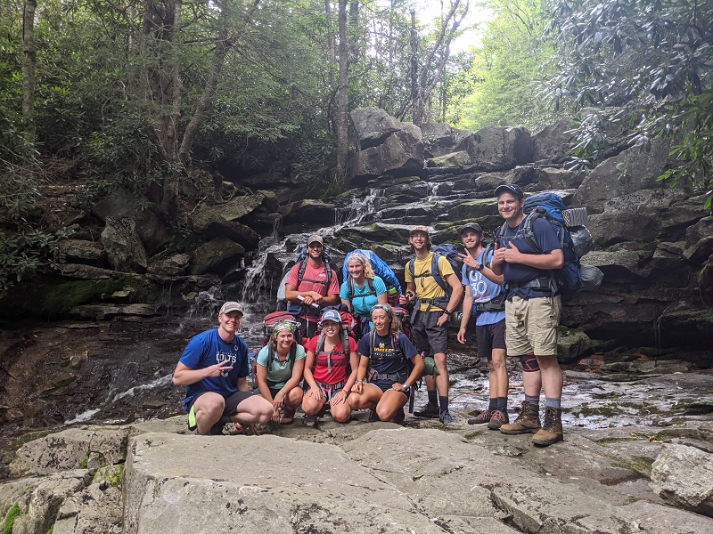${ Nine students smiling on an outdoor backpacking trip in front of a waterfall  }