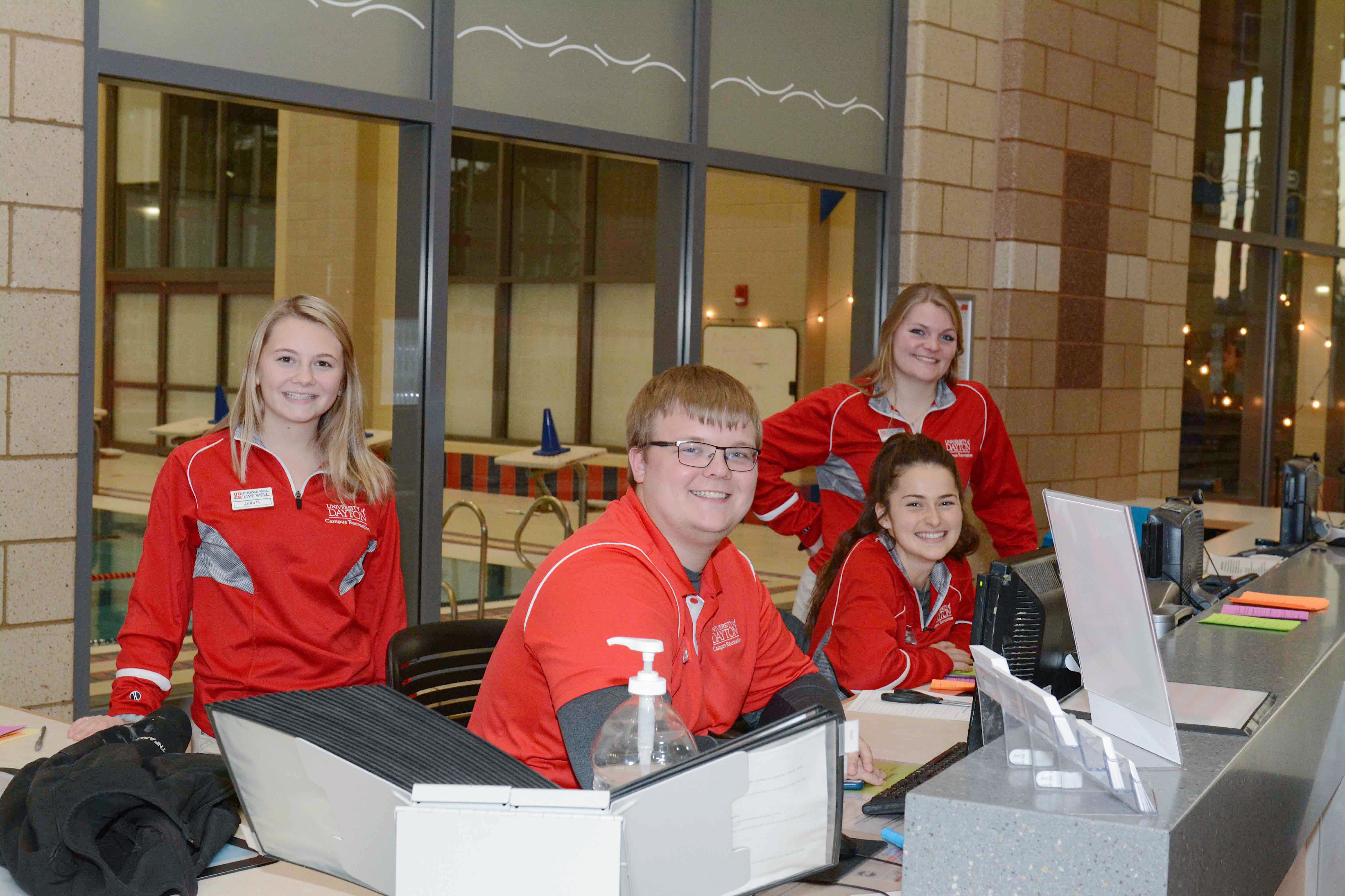 ${ Four facility operations attendants smiling at front desk of the RecPlex. }