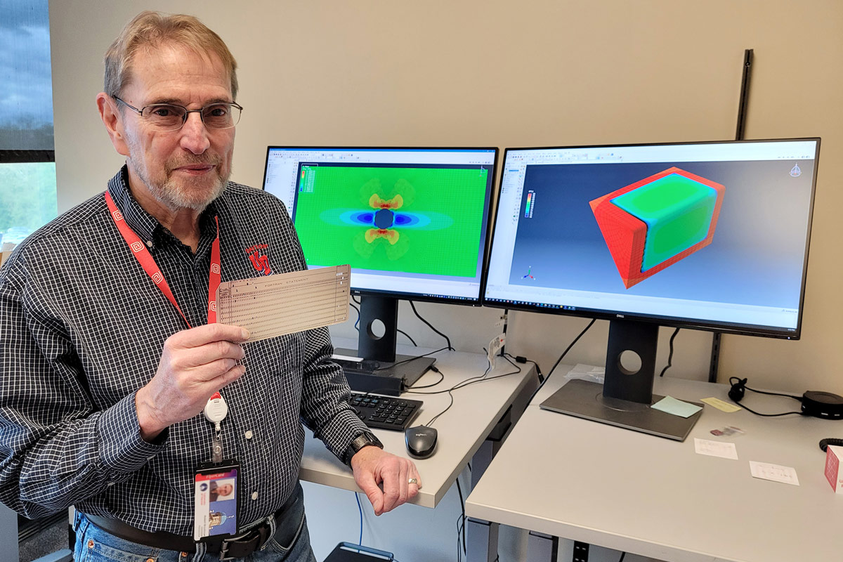 Bob Brockman holds a replica of a Fortran punch card while standing next to two computer monitors.
